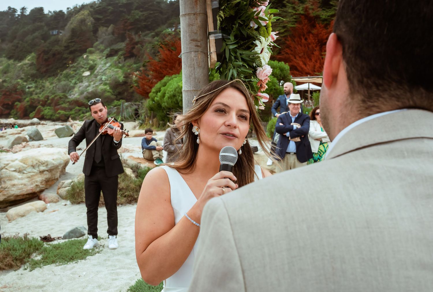 Bride speaks vows into microphone facing groom with violinist performing in background at wedding.