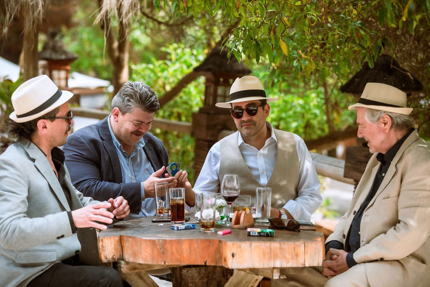 Four men in hats enjoying cigars and drinks at a wooden outdoor table under shaded trees.