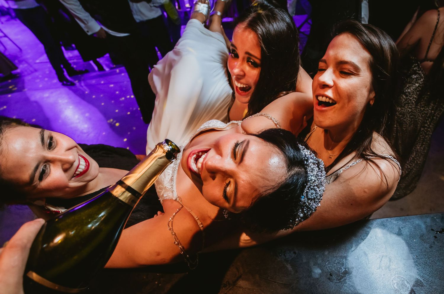 Group of women laugh together holding champagne bottle at festive wedding celebration.