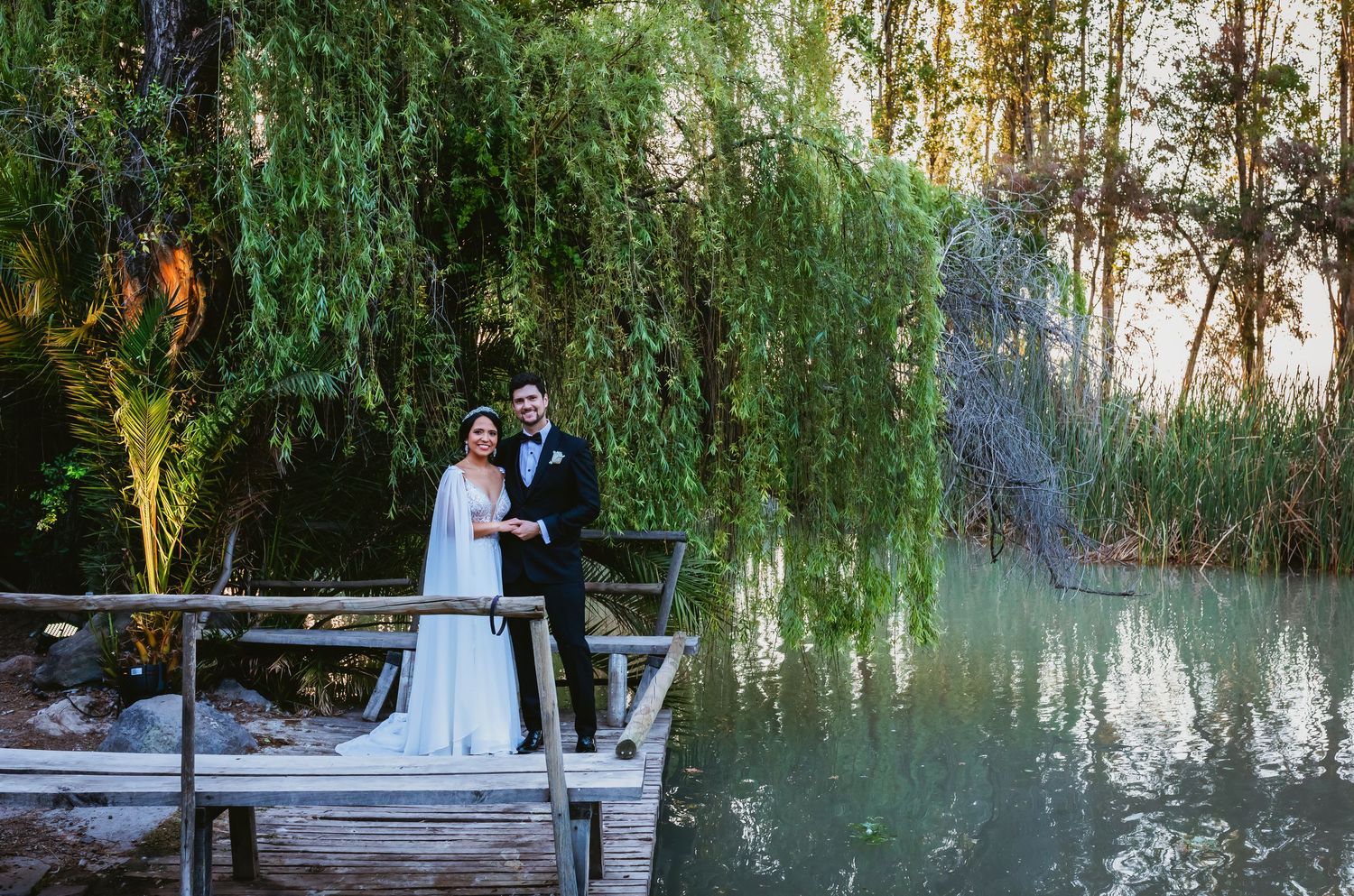 Wedding couple holds hands on wooden dock surrounded by weeping willow trees and tranquil green water.