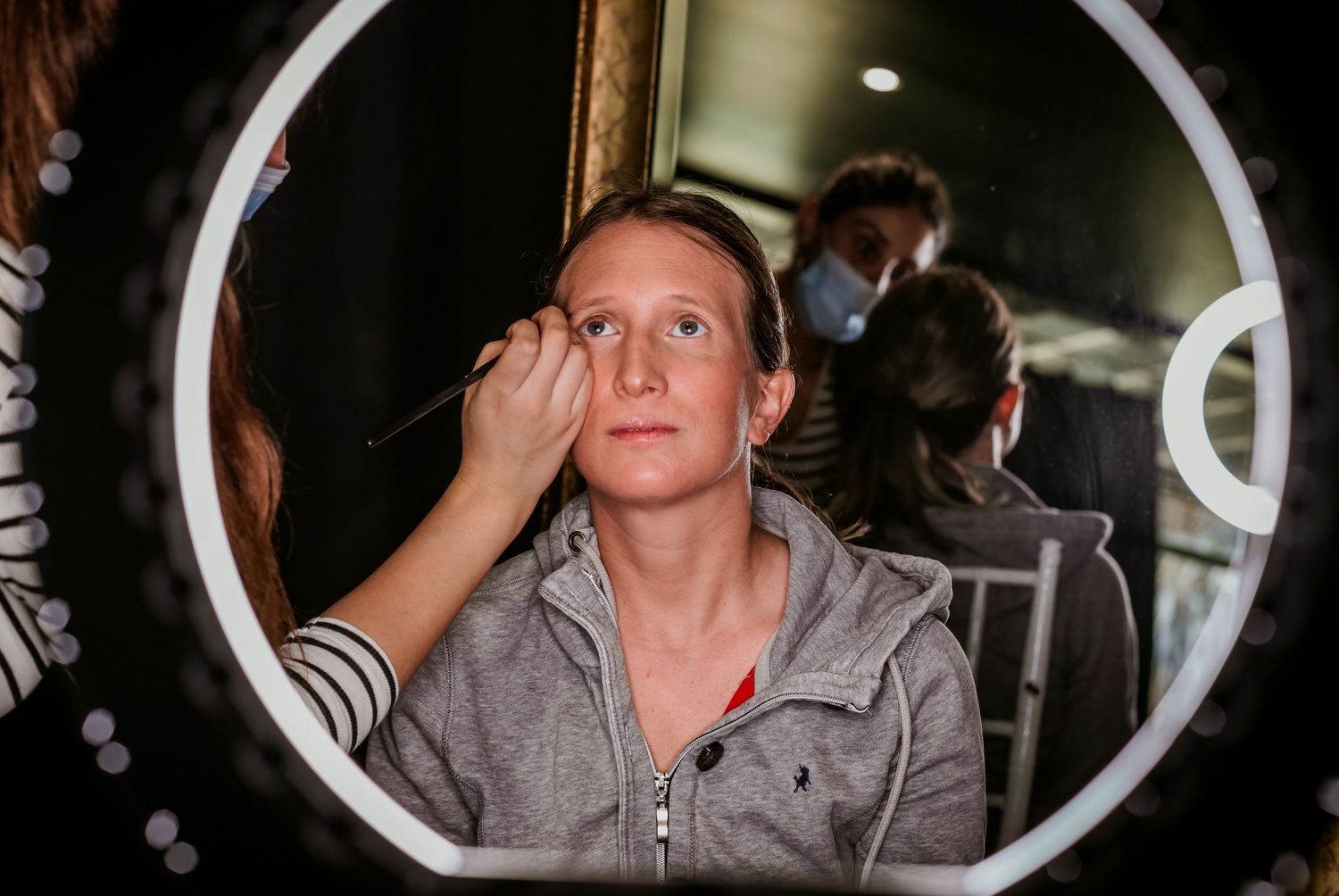 A makeup artist applies eyebrow makeup to a woman reflected in a round LED mirror.