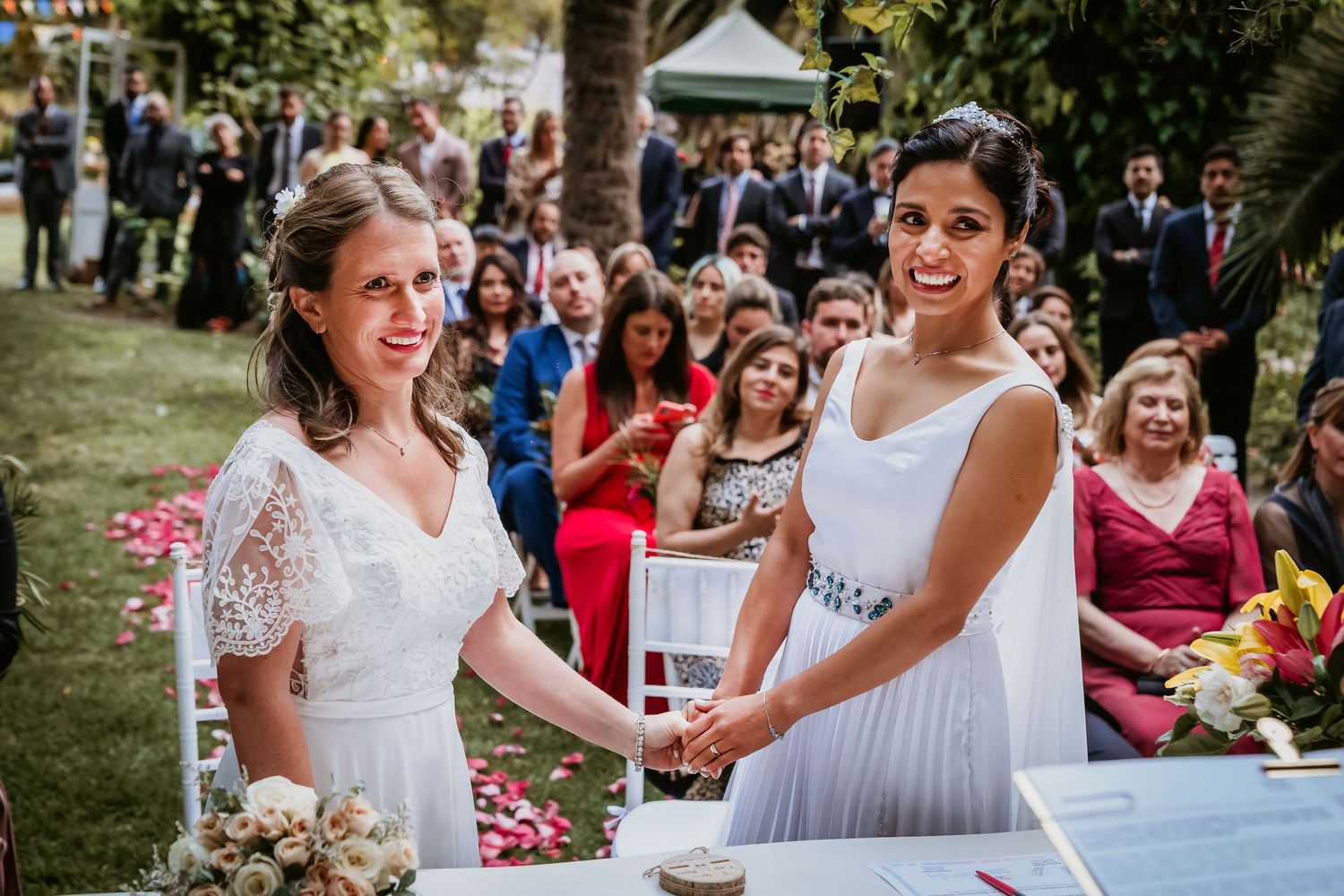 Two brides hold hands and smile at a signing table during their outdoor wedding.