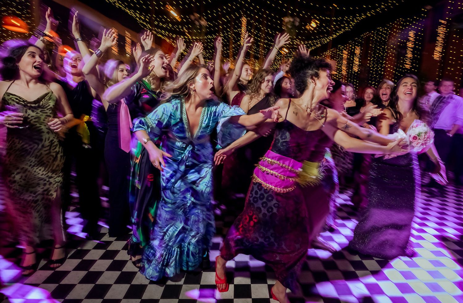 Guests in colorful formal gowns dancing energetically on checkered dance floor at wedding reception.