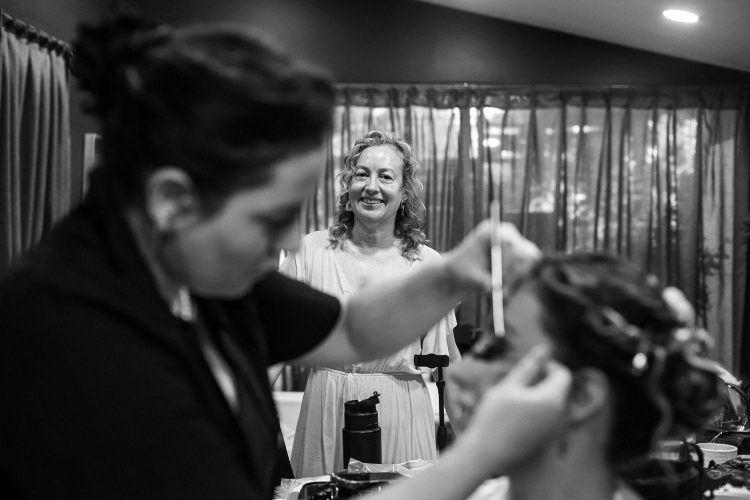 Black and white photo of bride smiling as makeup artist works on bridesmaid during wedding prep.