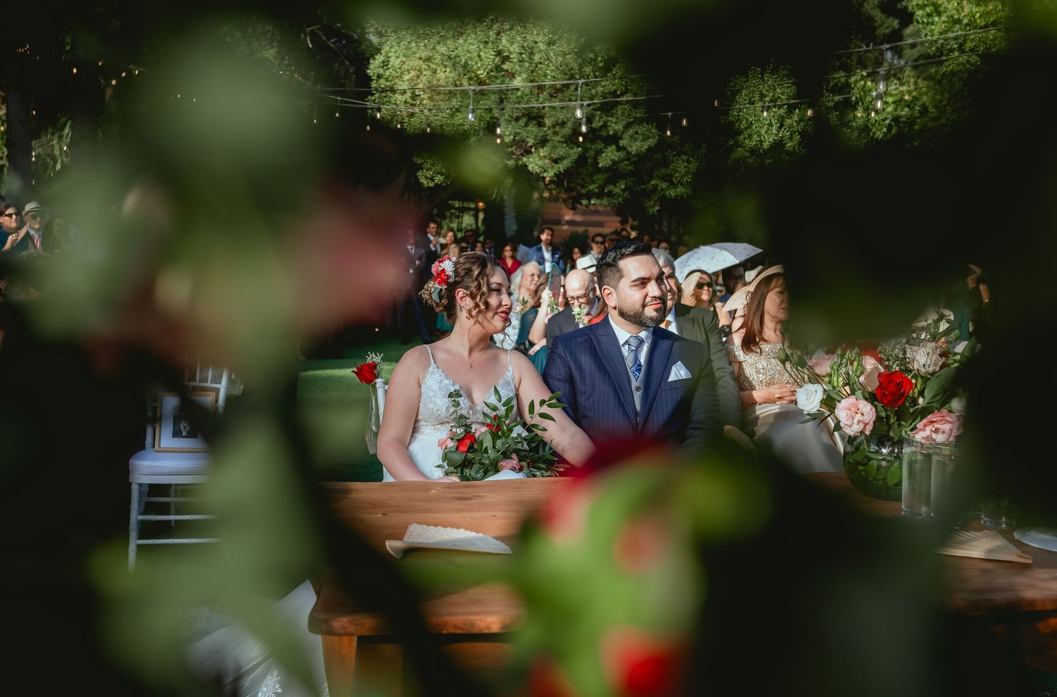 Bride and groom share a smile at outdoor wedding ceremony table, framed by colorful floral bokeh.