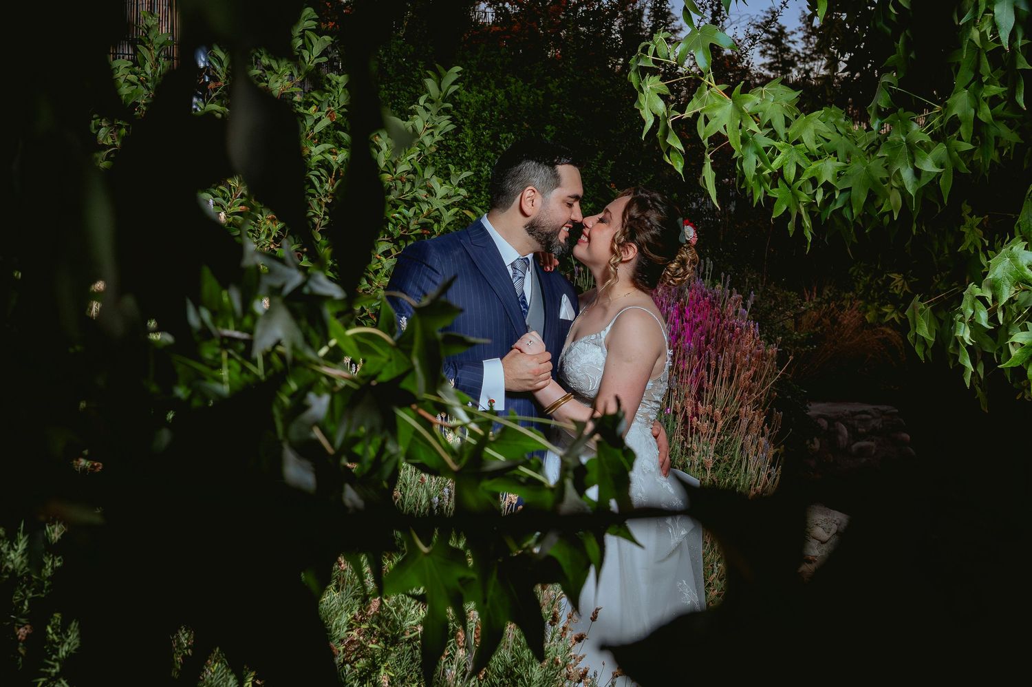 Bride and groom share a gentle kiss framed by vibrant green leaves in garden.