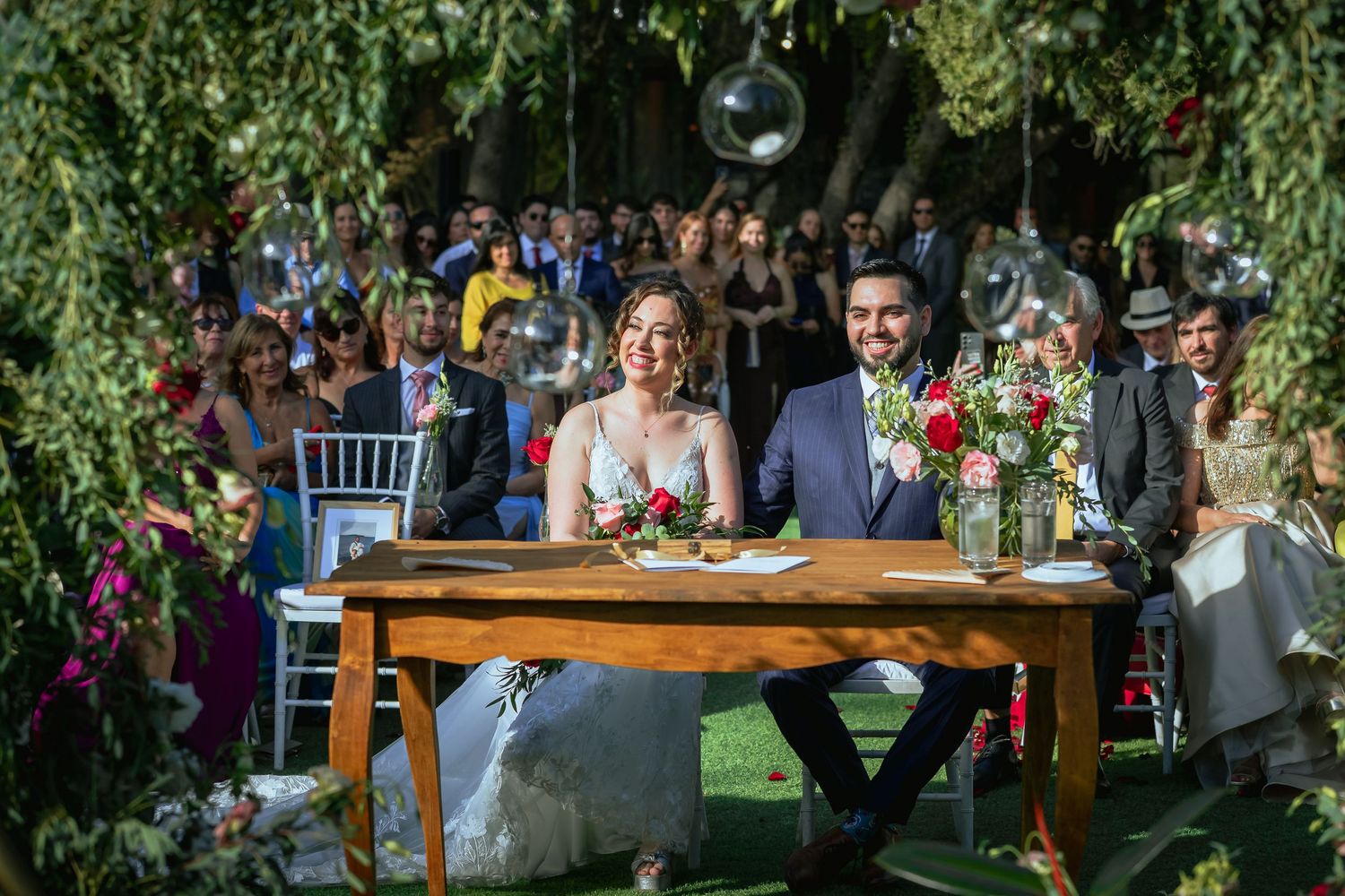 Smiling bride and groom sit at signing table during outdoor wedding ceremony under green trees.