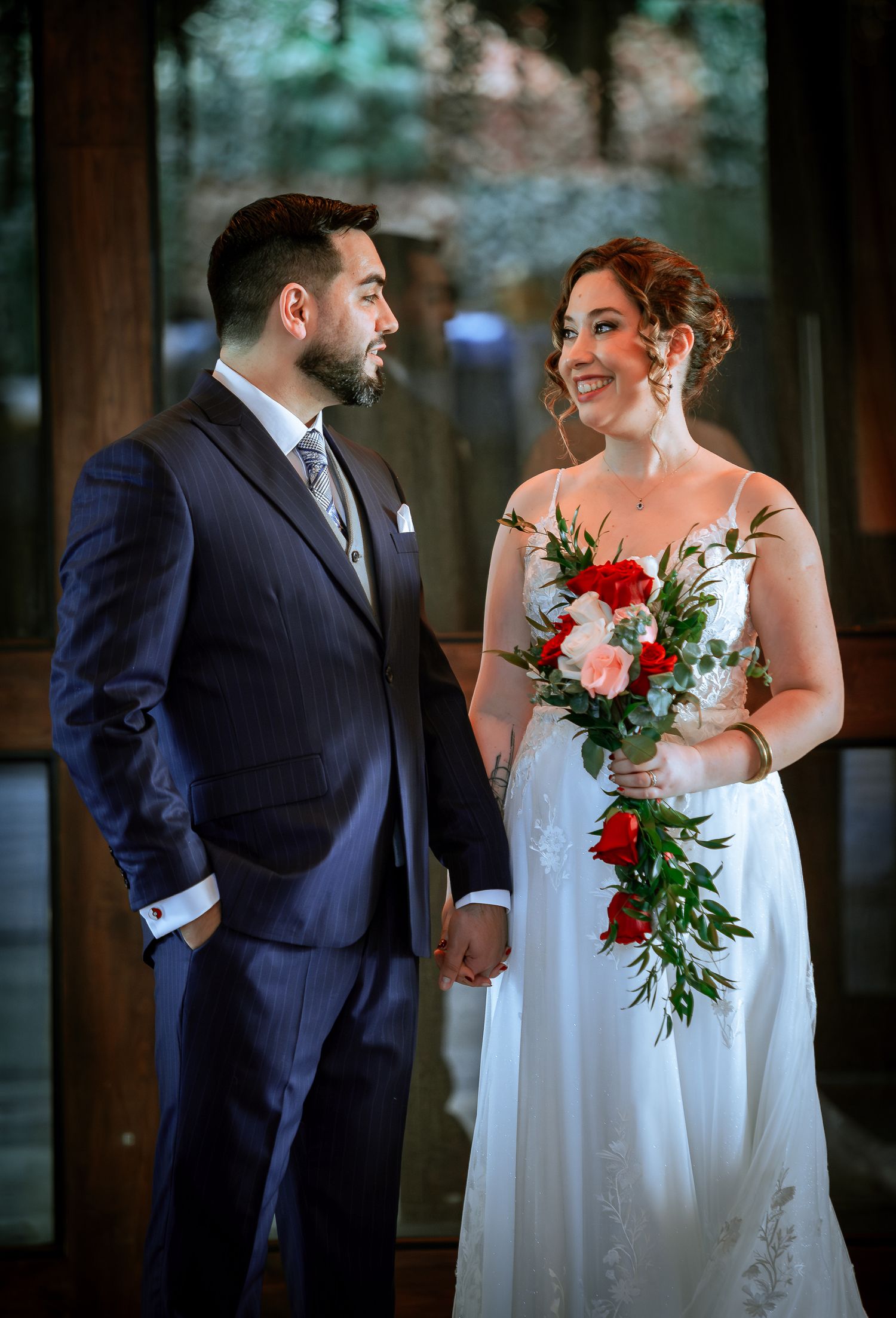 Bride holding a red and pink rose bouquet stands with groom at their outdoor wedding venue.