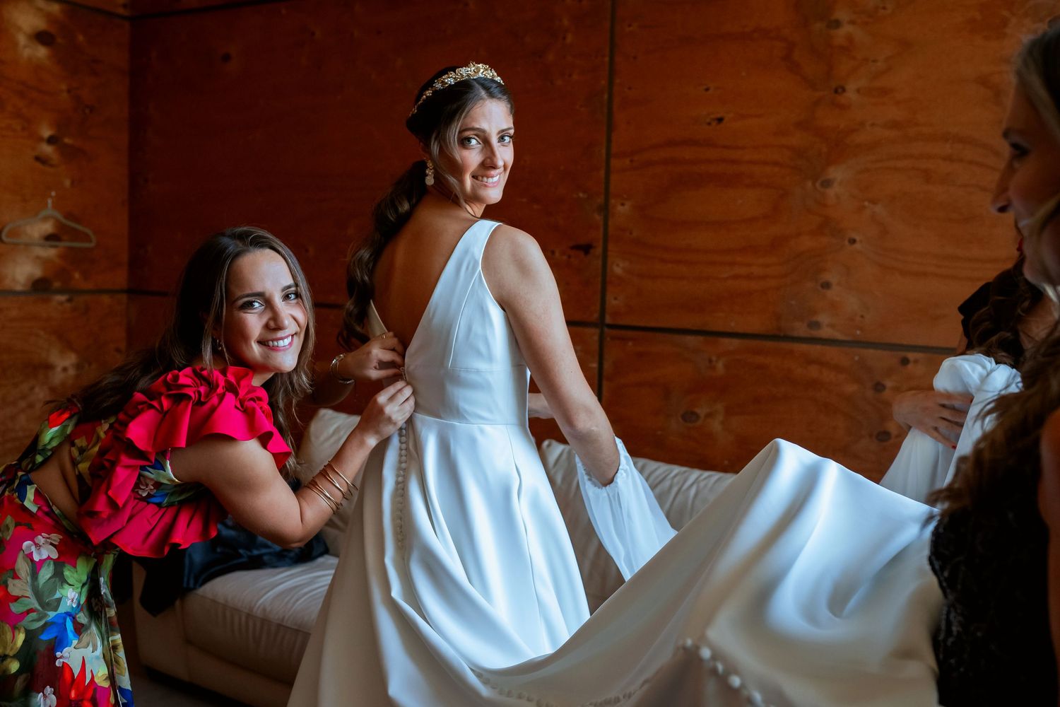 Bridesmaid in floral dress helping bride button her elegant white wedding gown before the ceremony.