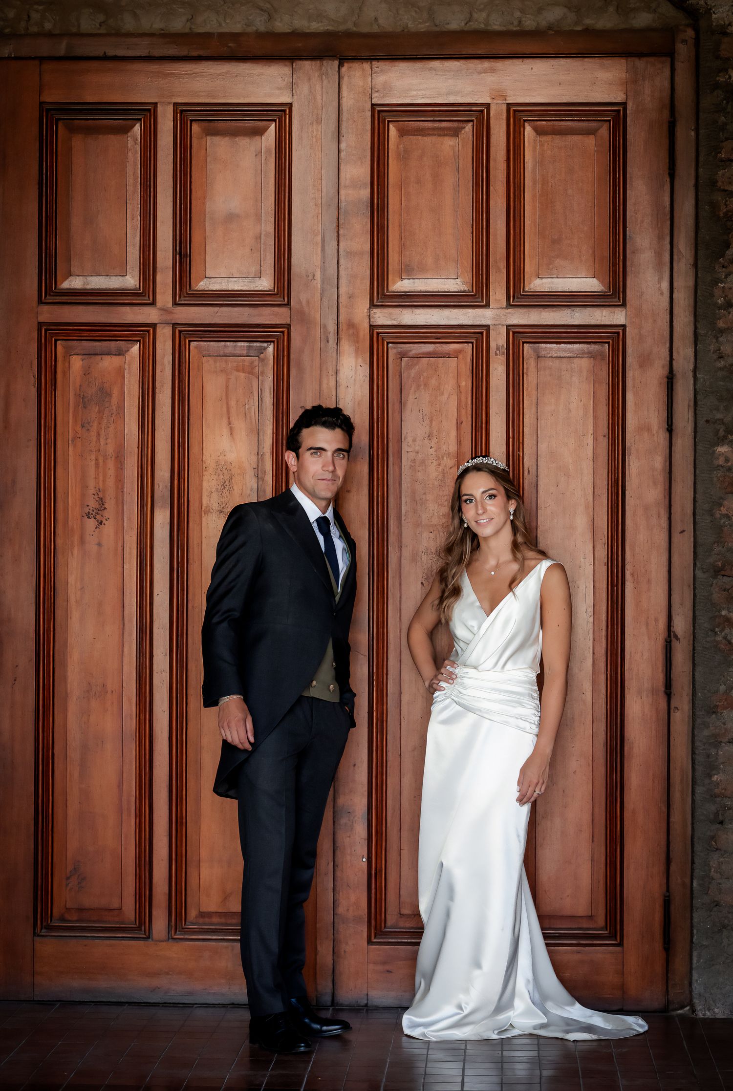 Bride in white gown and groom in dark suit posing before large ornate wooden doors.