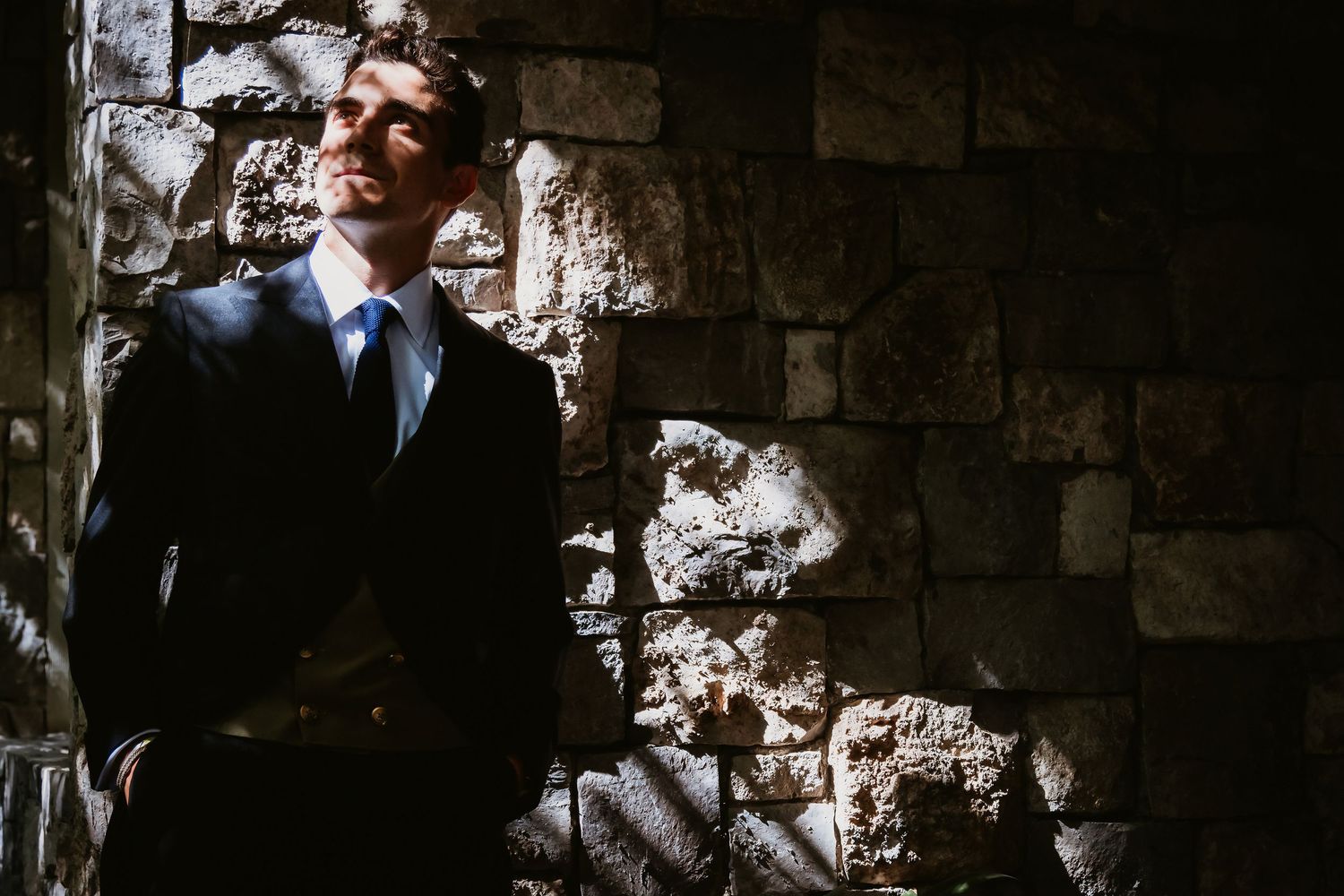Groom in dark suit leaning against rustic stone wall in dramatic light and shadow.