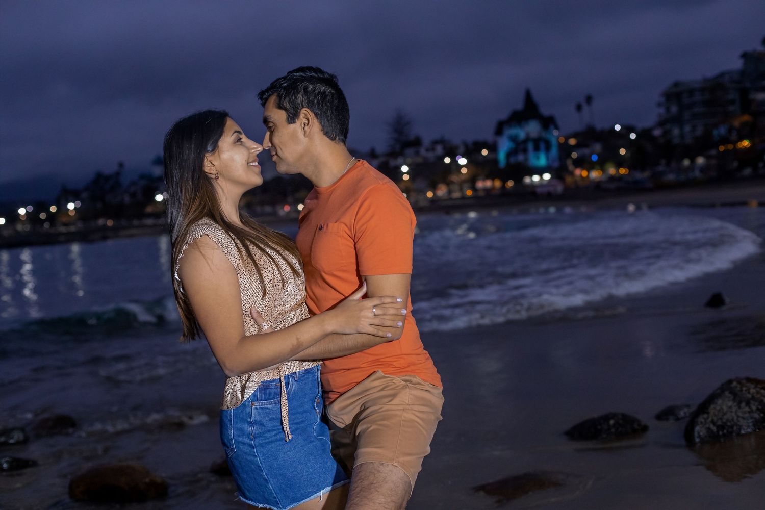 Couple embracing on a sandy beach at twilight with illuminated buildings in the background.