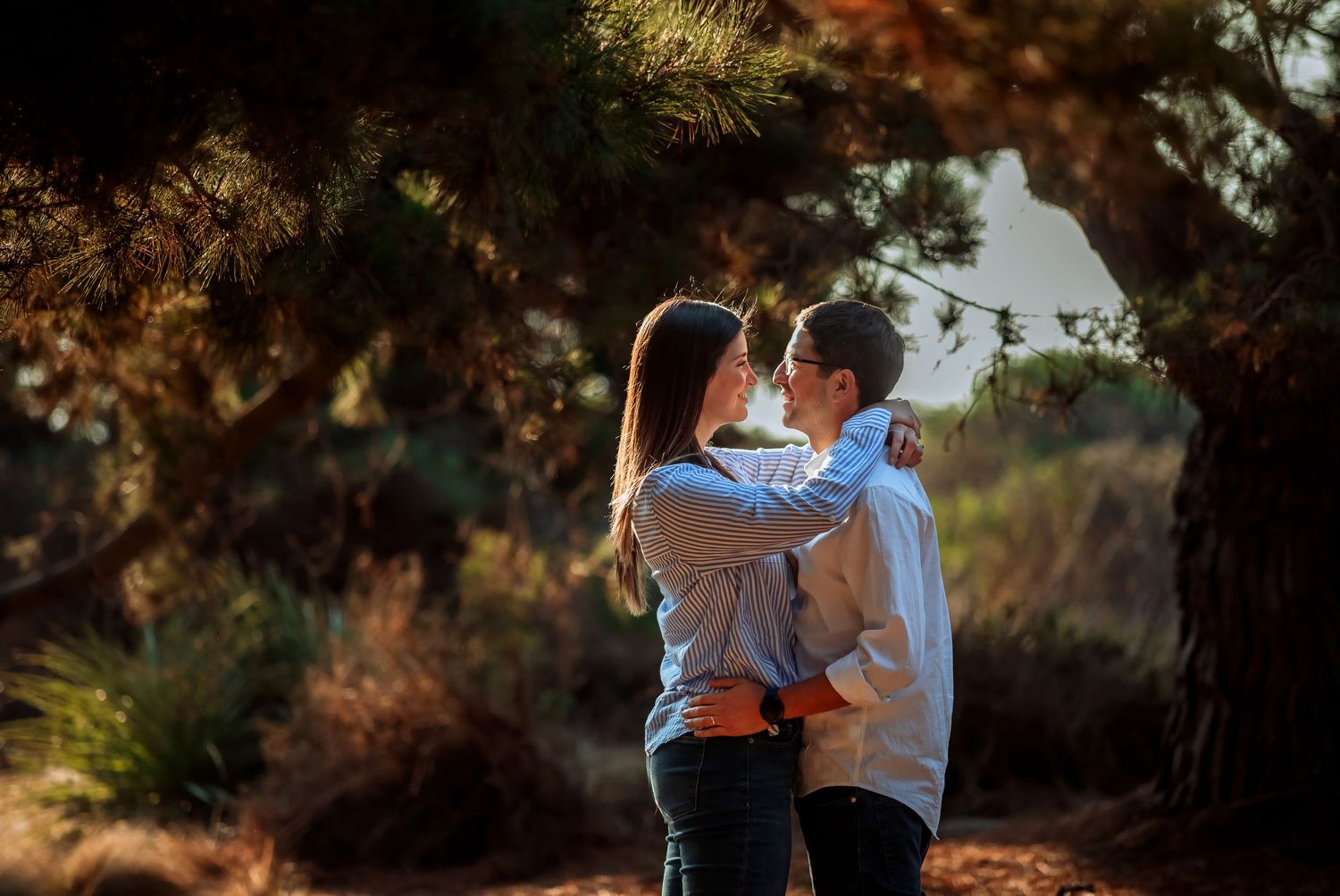 Romantic couple sharing a kiss under pine trees in warm sunset light.