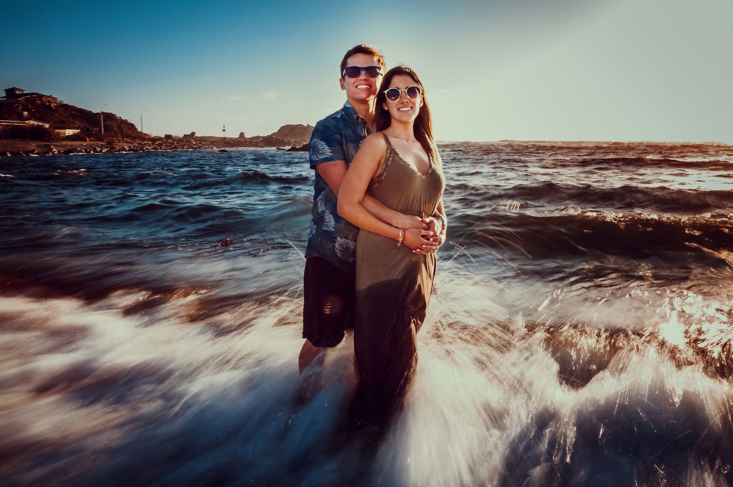Couple embracing in ocean waves with a coastal town visible in the background.
