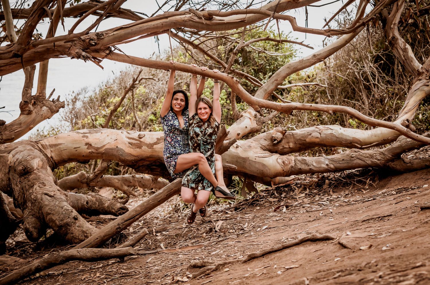 Two women in floral dresses posing together under arching driftwood branches on a trail.