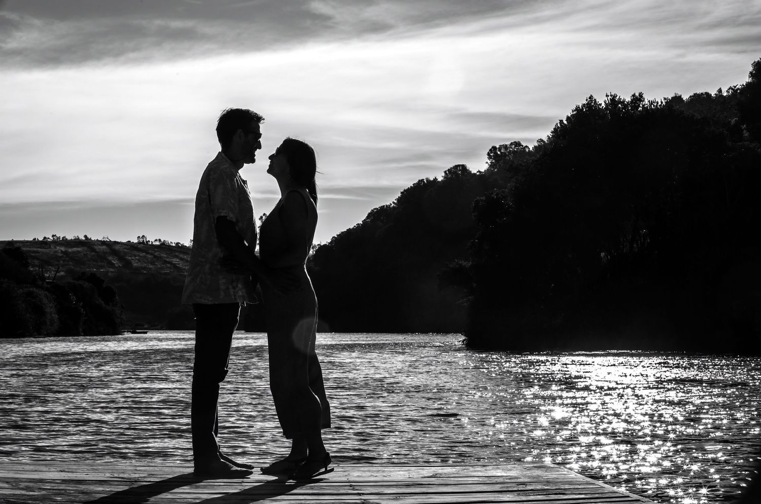 Black and white silhouette of a couple standing by a shimmering river at sunset.