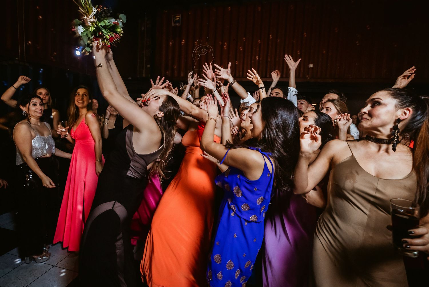 Excited women reaching up to catch a bridal bouquet toss on a dark wedding reception dance floor.