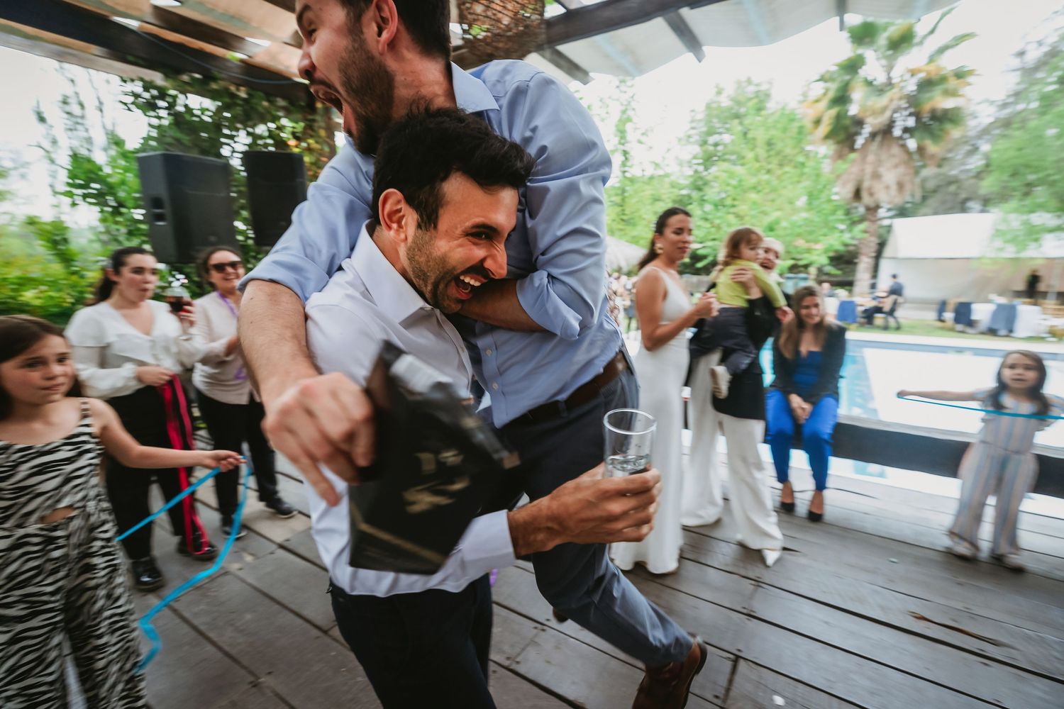 Man being lifted by friend at lively outdoor wedding celebration on wooden deck with guests watching.