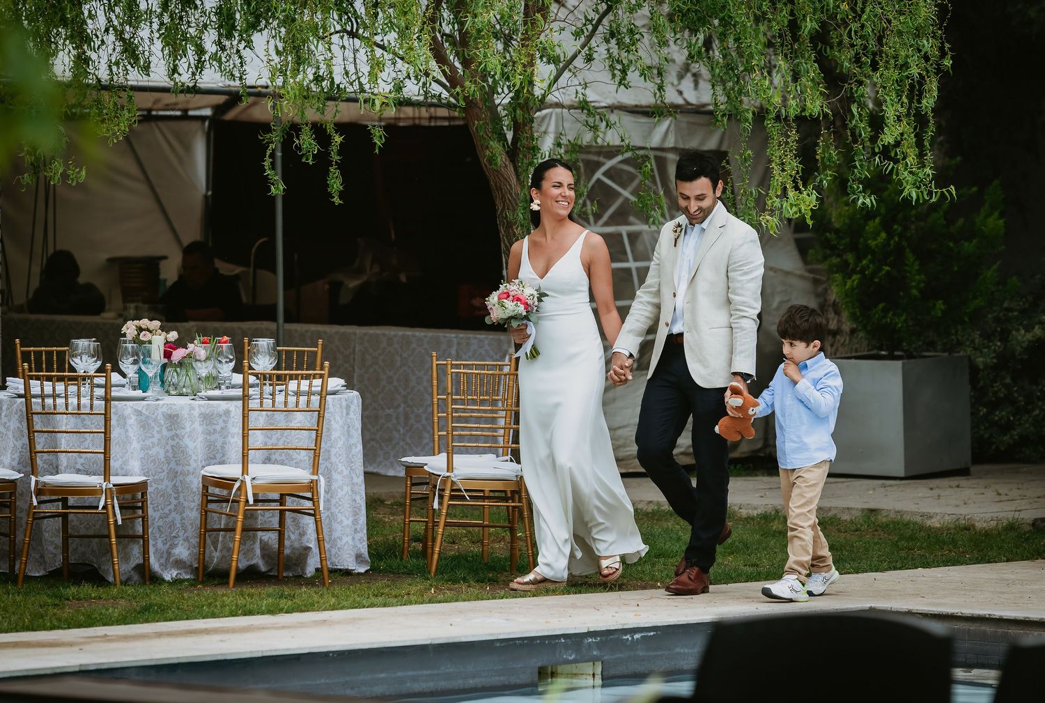 Bride and groom walk hand-in-hand with a young boy at an outdoor wedding reception near a pool.