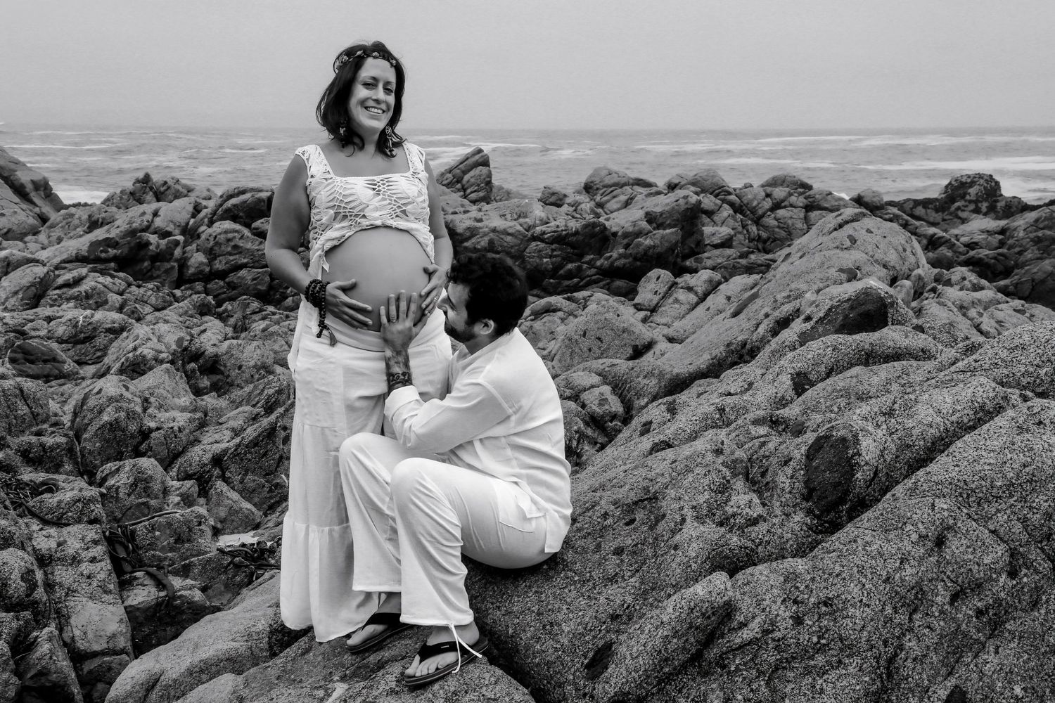 Black and white maternity photo of couple on rocky beach, woman standing showing baby bump, man seated beside her.