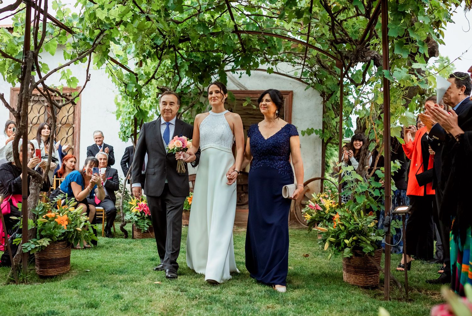 Bride in silver gown walks down garden aisle escorted by parents under romantic vine-covered wedding arch.