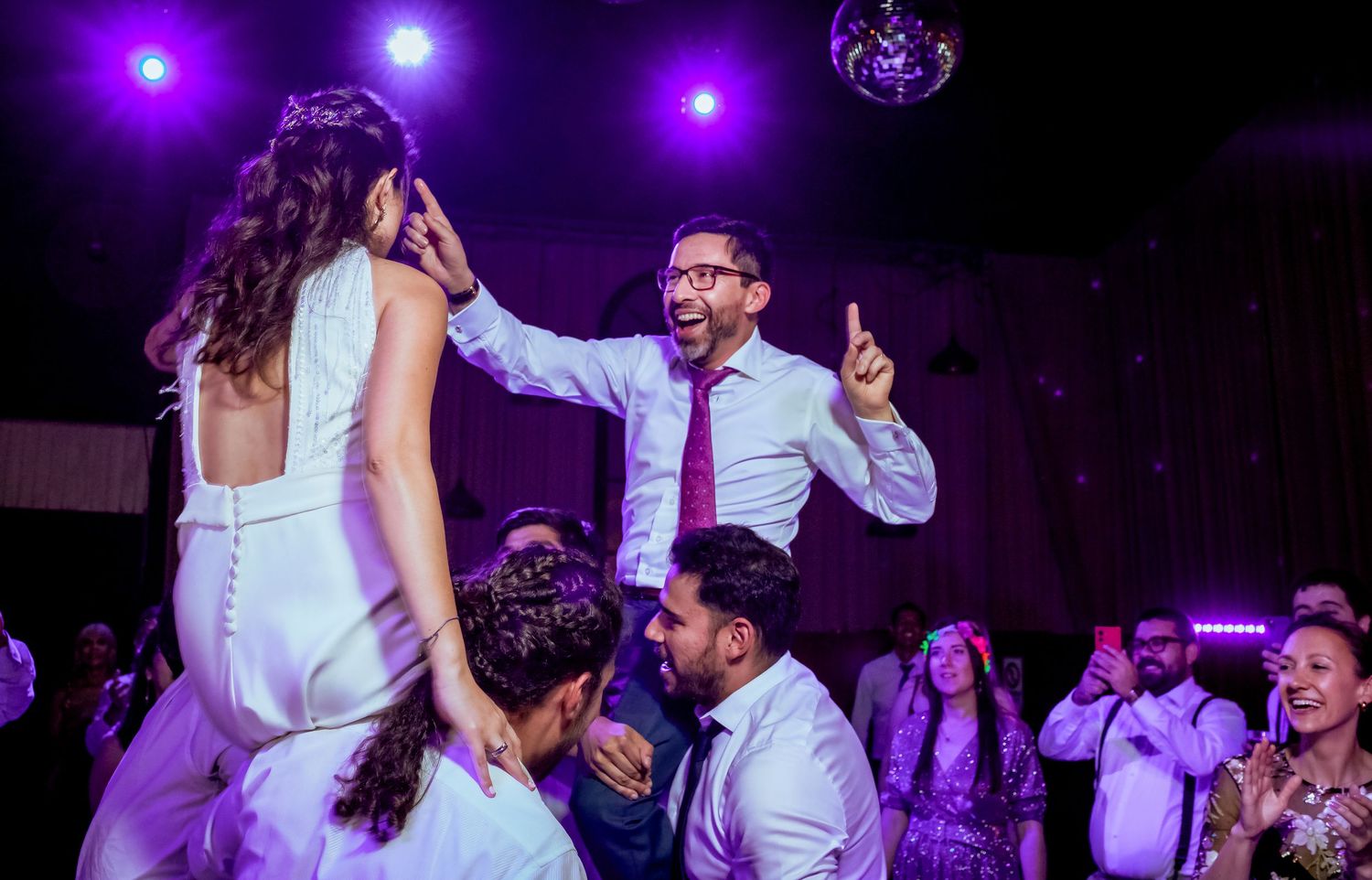 Groom lifts bride on dance floor at lively wedding reception with purple lights.