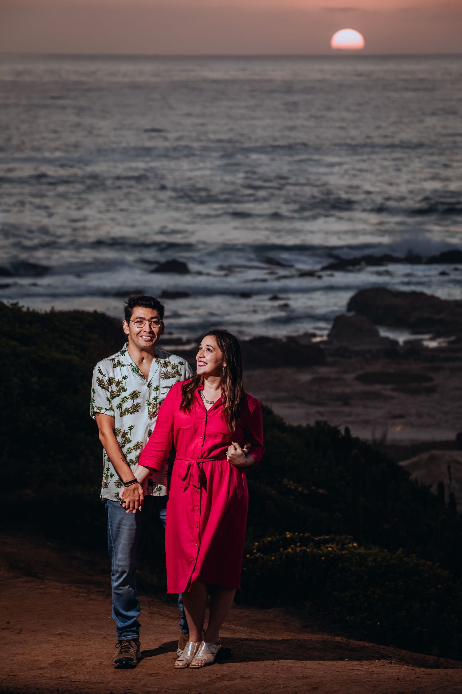 Couple holding hands walking along a coastal path at sunset with ocean waves and glowing sun in the background.