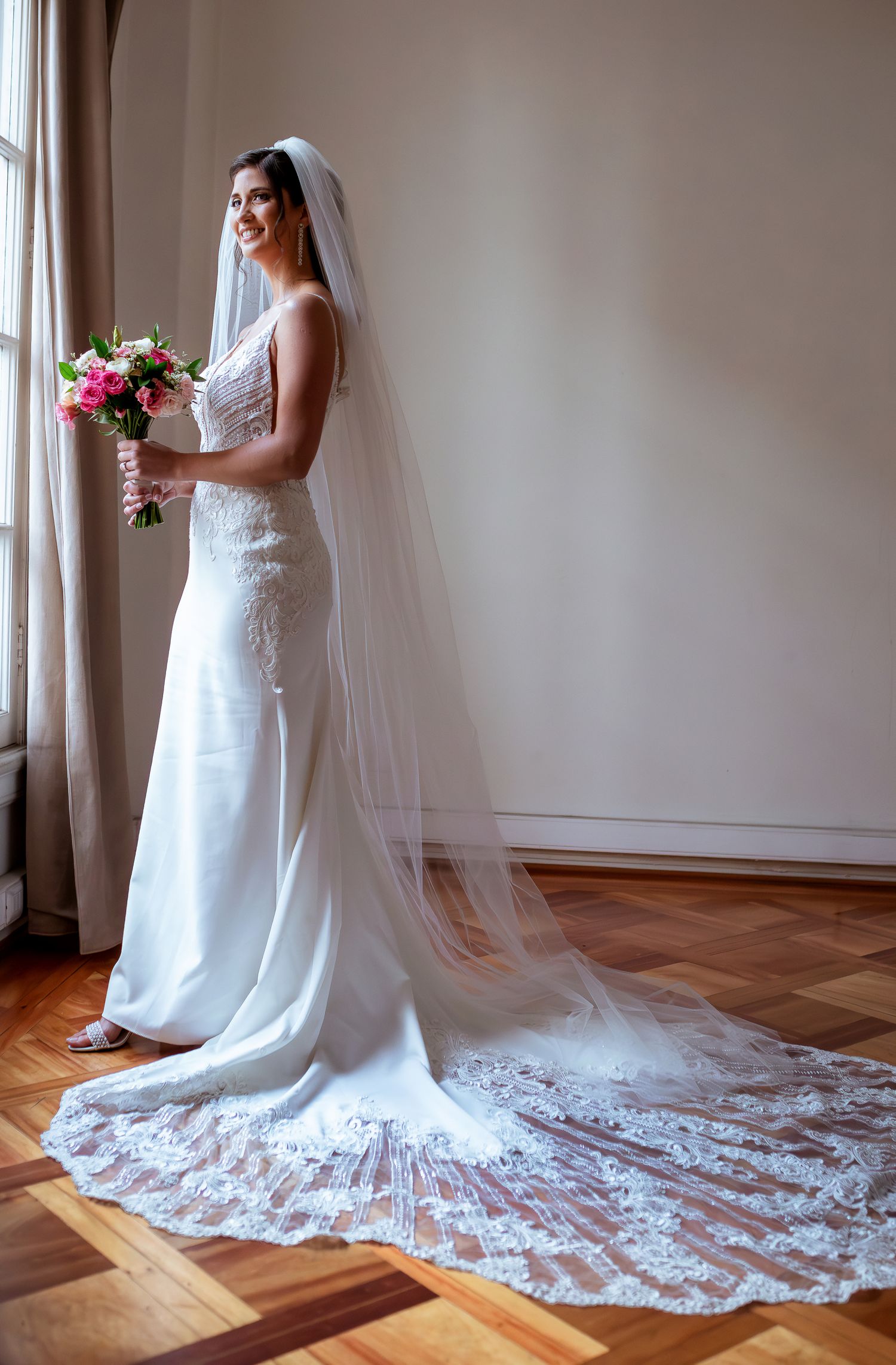 Bride in white fitted wedding dress with long lace veil poses elegantly by bright window.