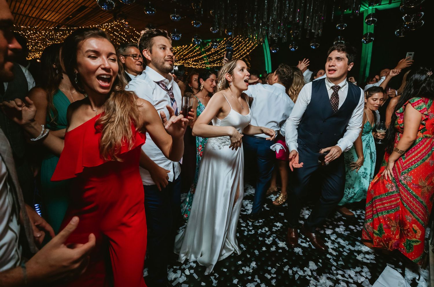 A joyful wedding reception with bride in white gown and guests dancing and celebrating on a confetti-filled dance floor.