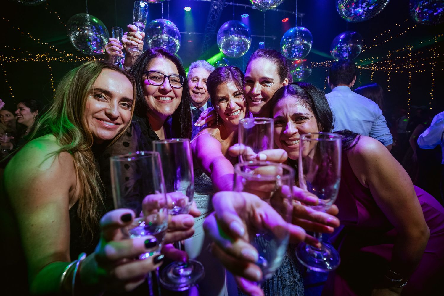 Group of women celebrating at a nightclub, raising champagne glasses under disco balls with colorful purple and green lights.