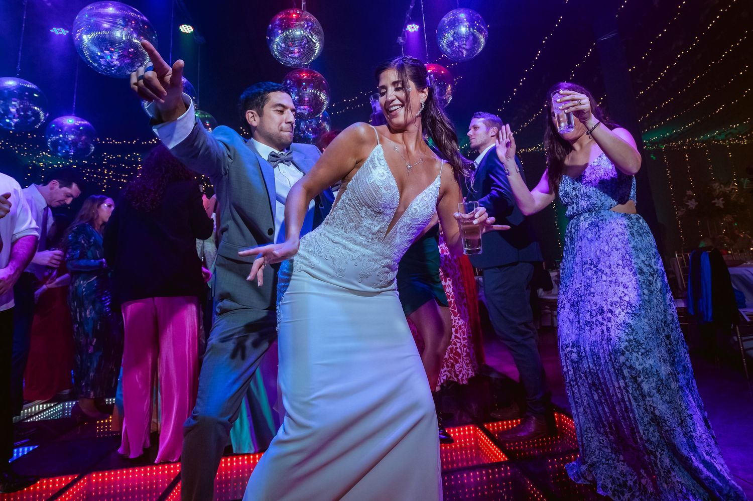 Bride in white gown dancing joyfully with guests under disco balls at a vibrant wedding reception.