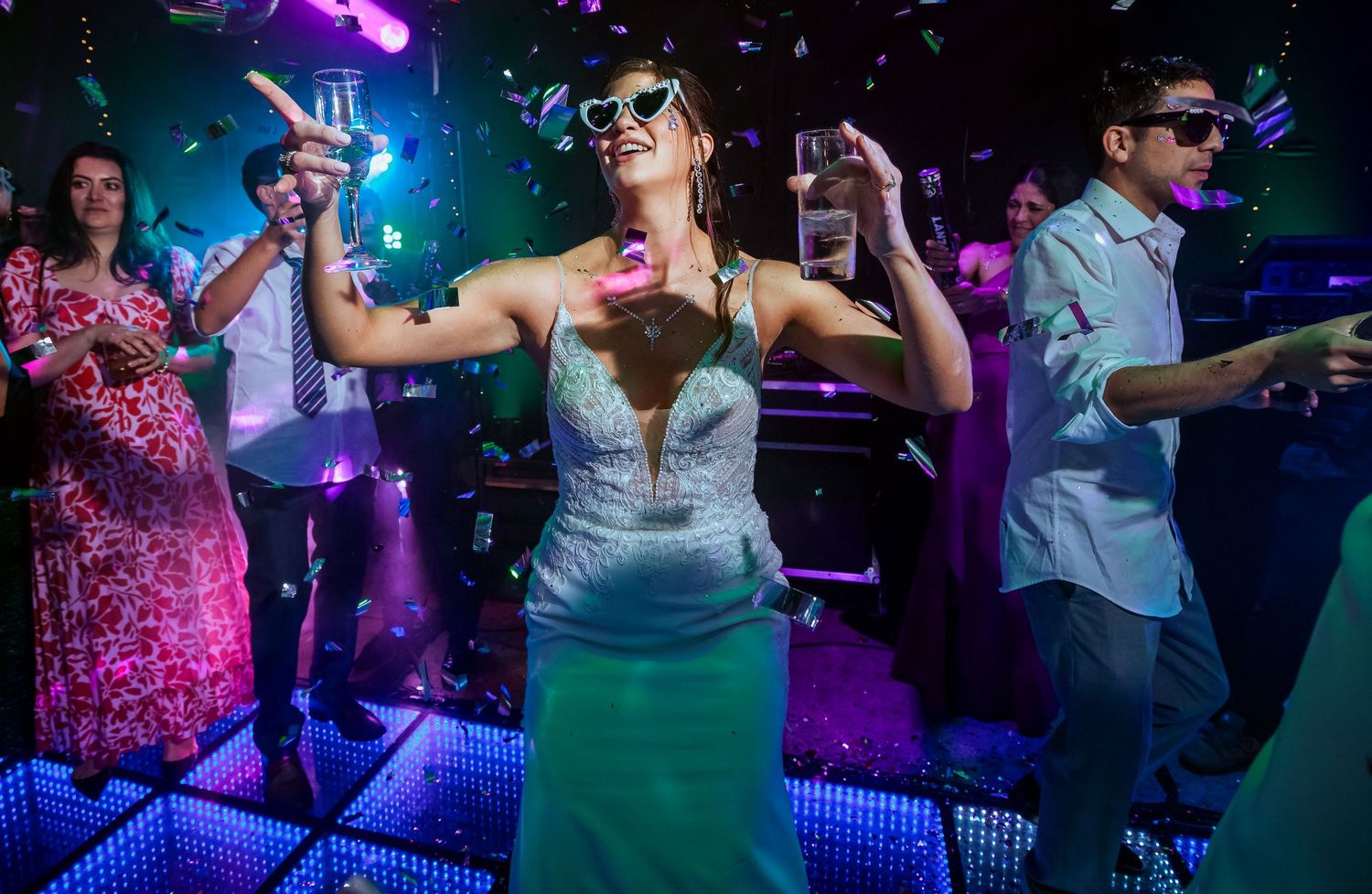 Woman in teal gown dancing joyfully at a vibrant party with confetti, colorful lights, and guests celebrating.