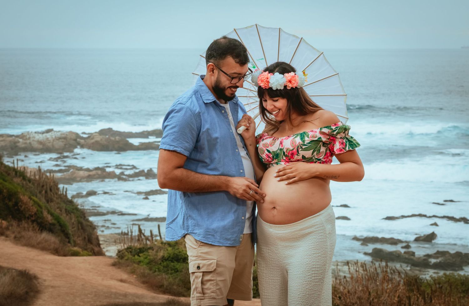 Couple smiling at pregnant belly outdoors by ocean with parasol and floral crown.