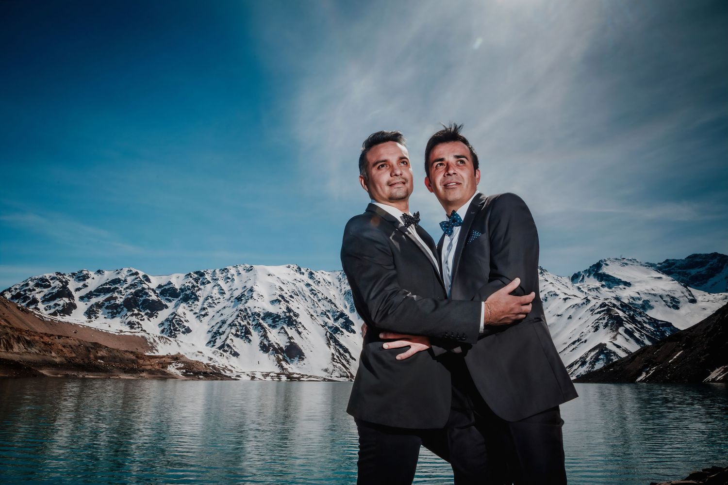 Same-sex couple embraces joyfully with snow-capped mountains and a blue alpine lake as backdrop.