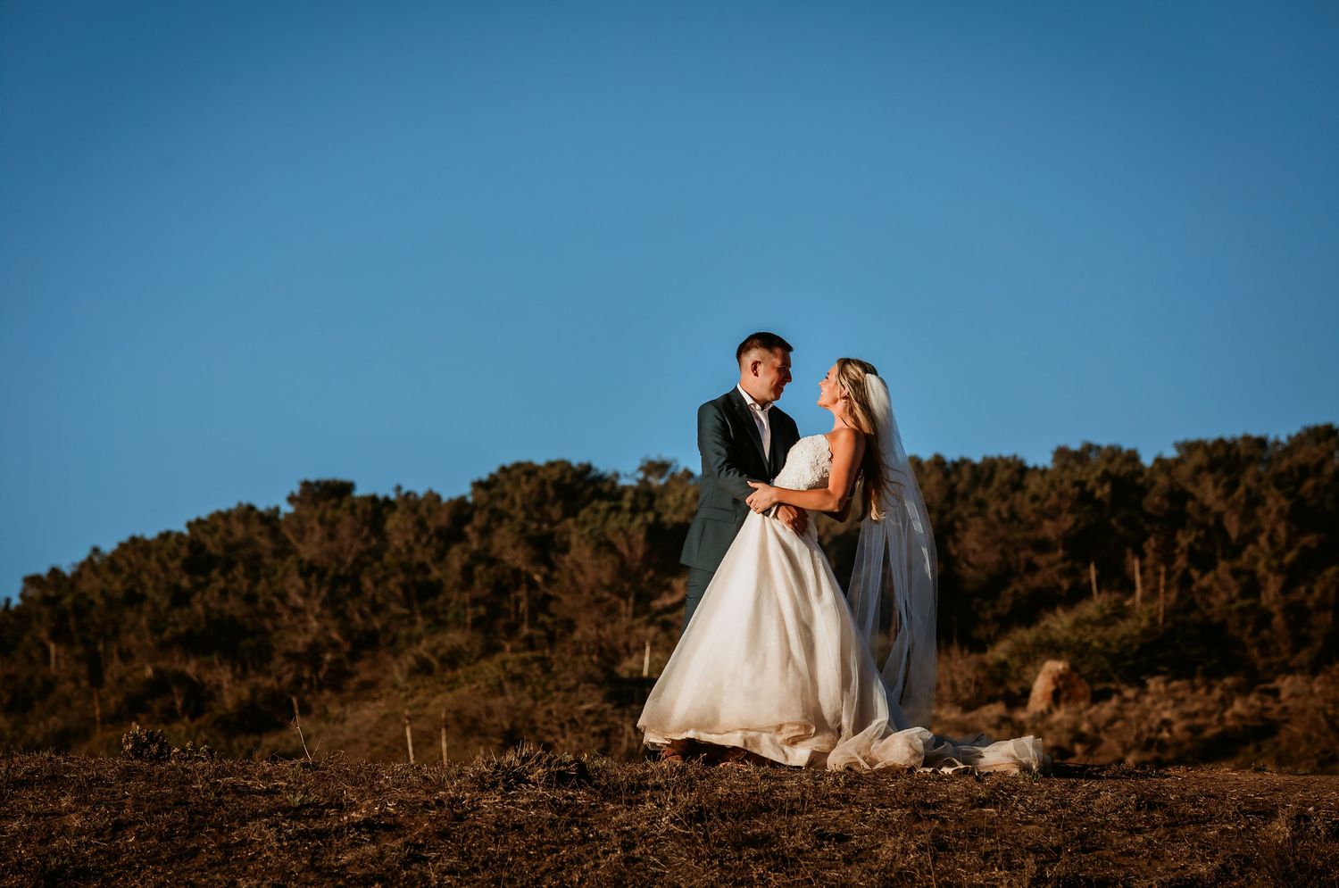 Bride and groom holding hands at golden hour on a hillside with blue sky backdrop.