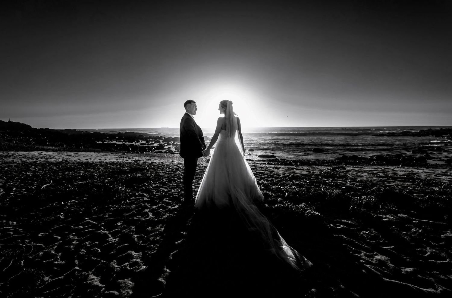Black and white silhouette of bride and groom holding hands on rocky beach at sunset.