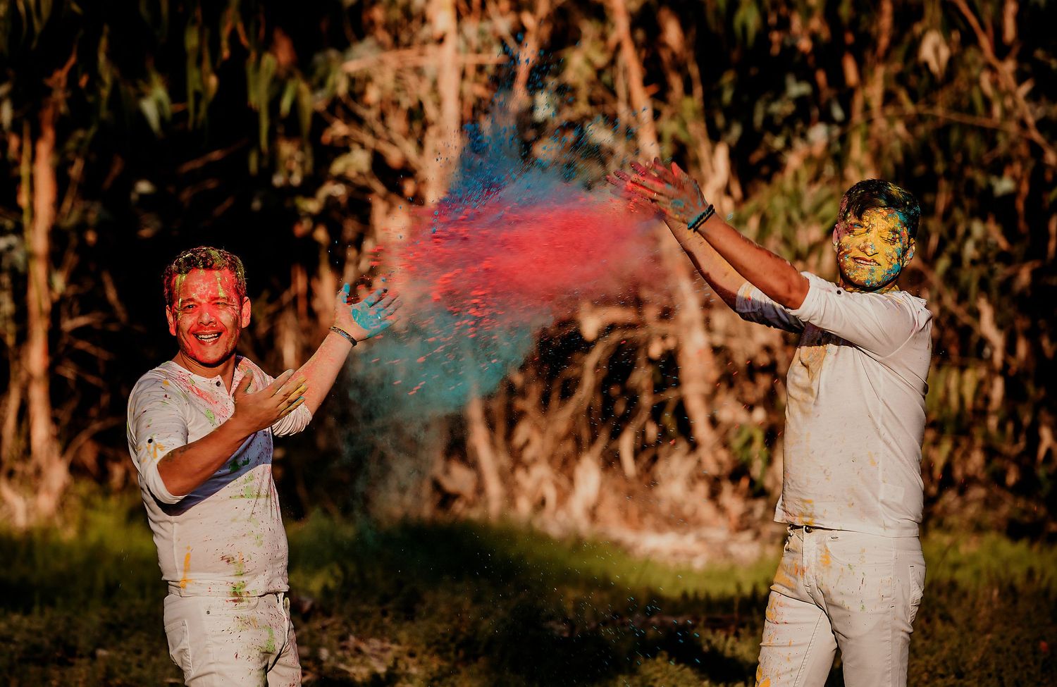 Two men joyfully throw red and blue Holi powder at each other outdoors near tall trees.