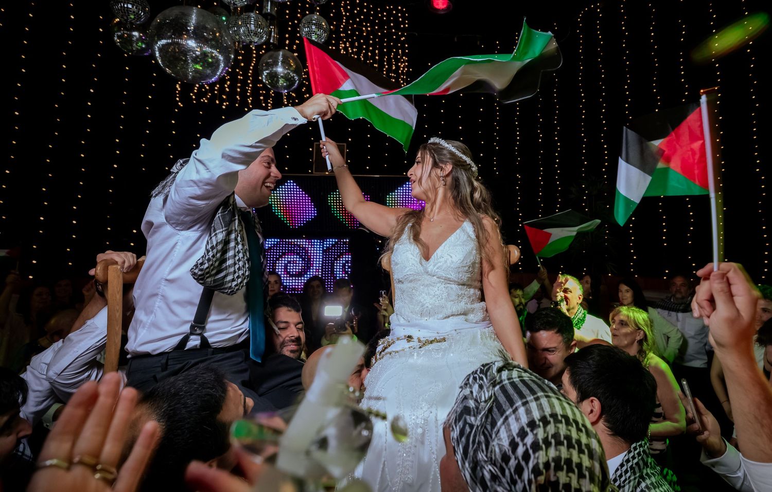 Bride and groom raised on chairs holding hands as guests wave Palestinian flags at a festive wedding reception.