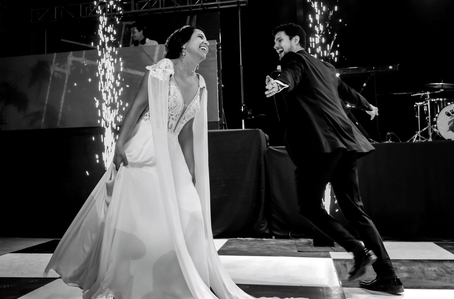 Bride and groom dancing joyfully on checkered floor with sparkling fireworks at elegant wedding reception.