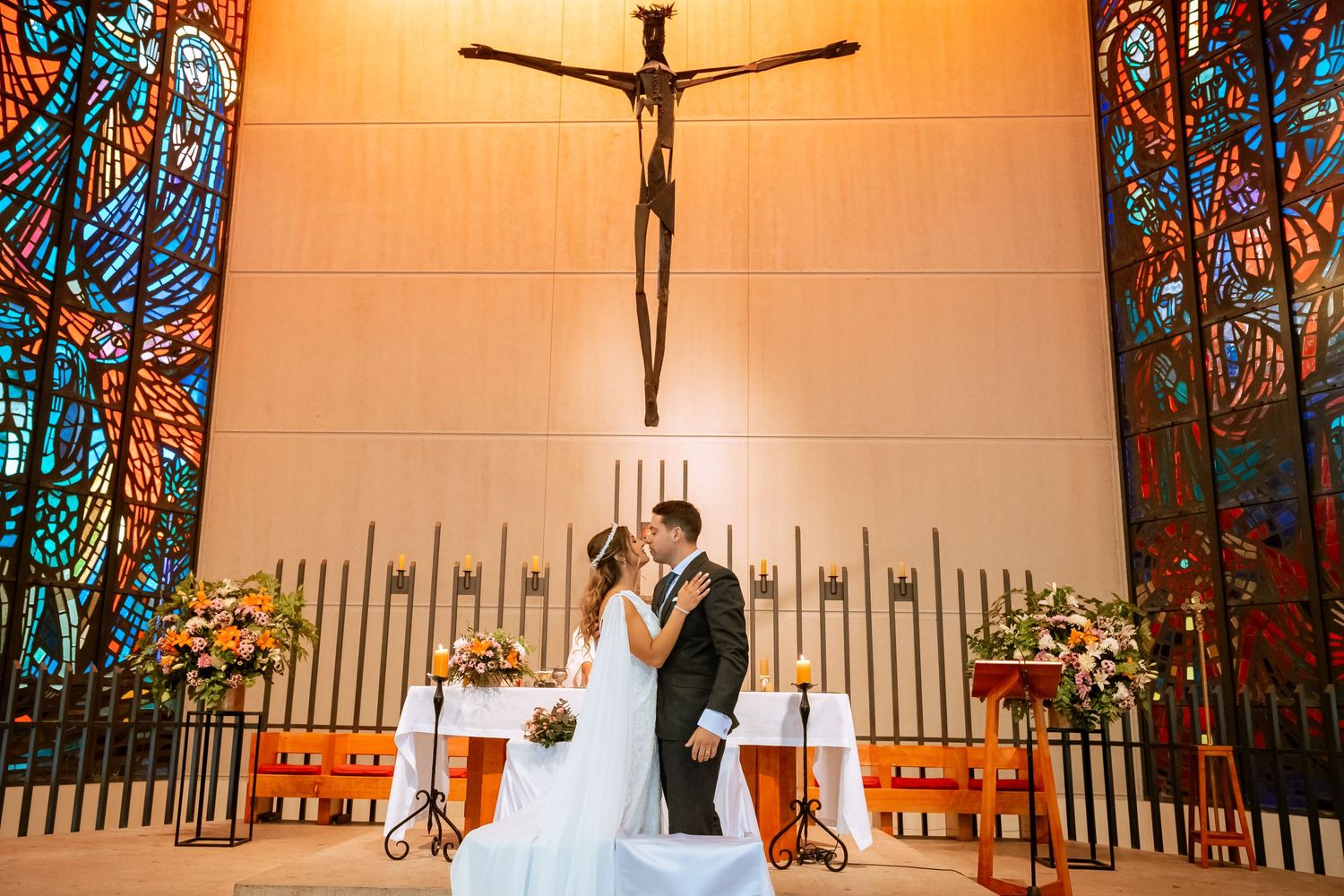 Newlyweds share a kiss at the altar beneath a crucifix with vibrant stained glass windows behind them.