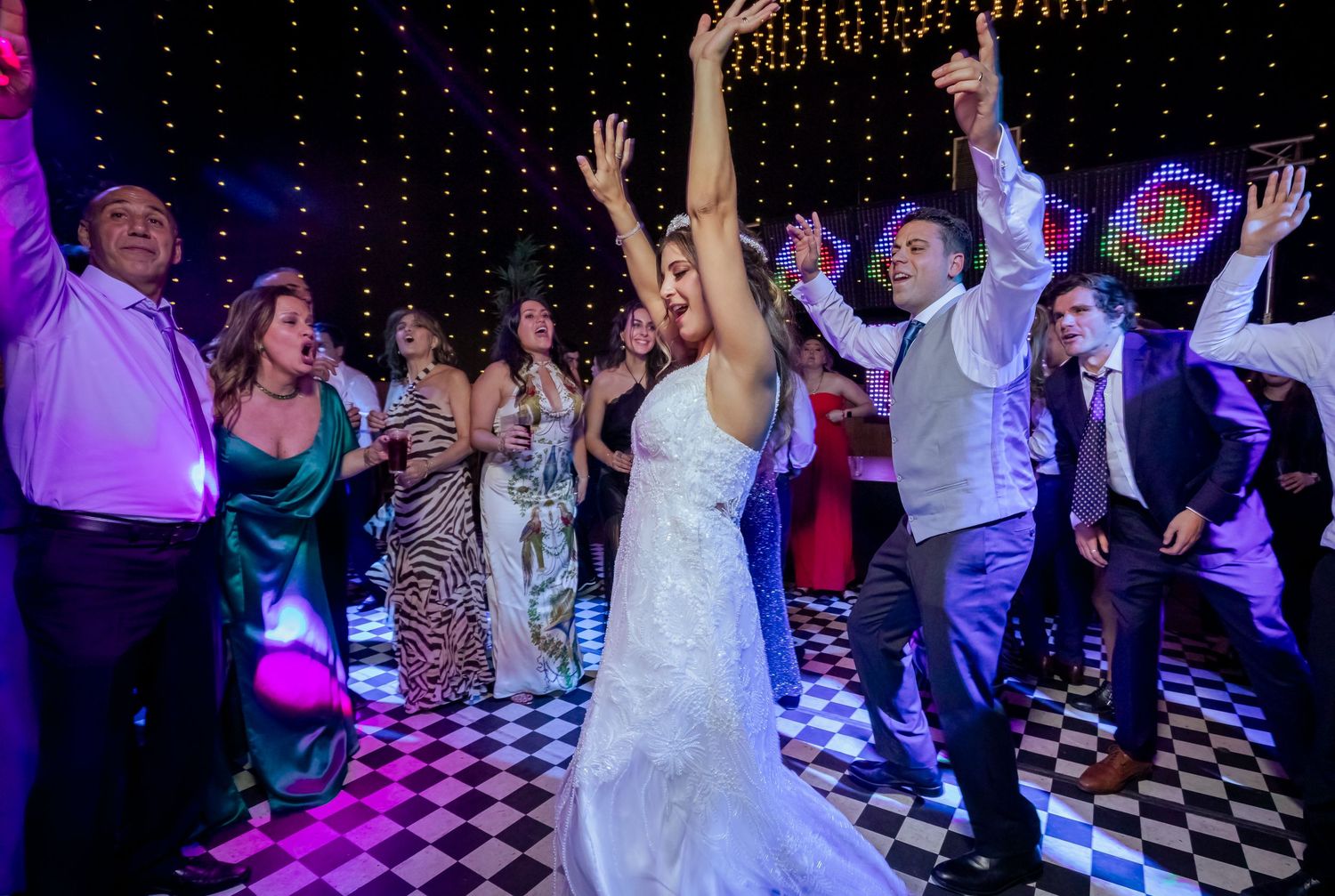 Bride raises her arms dancing with guests at an energetic wedding reception with string lights.
