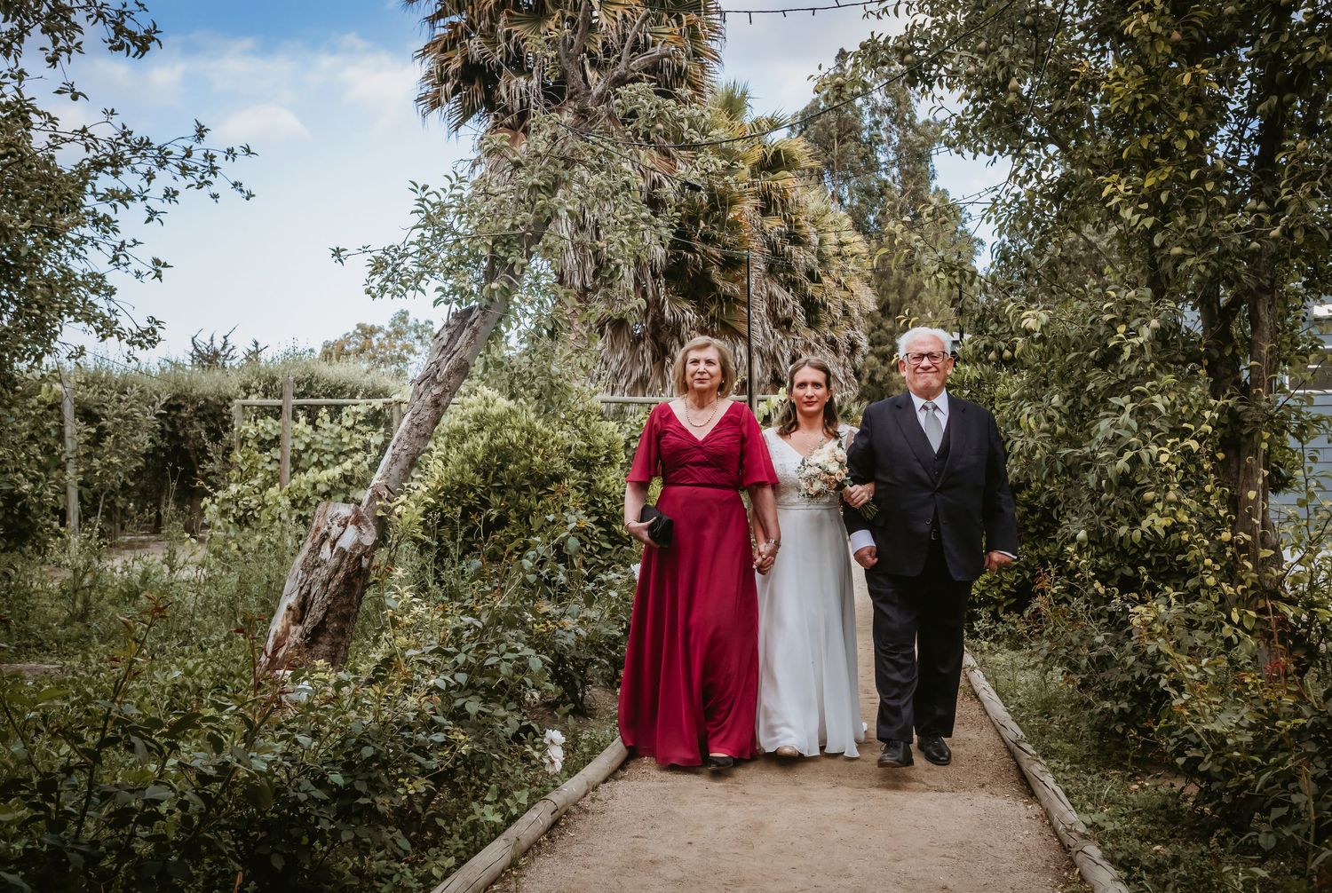 Bride walks garden path flanked by woman in red gown and man in dark suit at outdoor wedding.