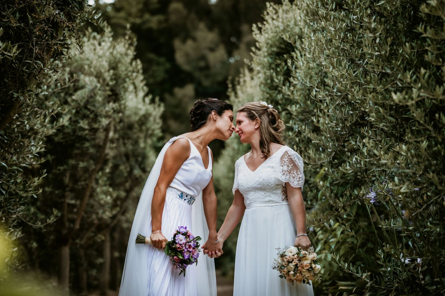 Two brides lean in for a kiss while holding bouquets along a lush olive tree pathway.