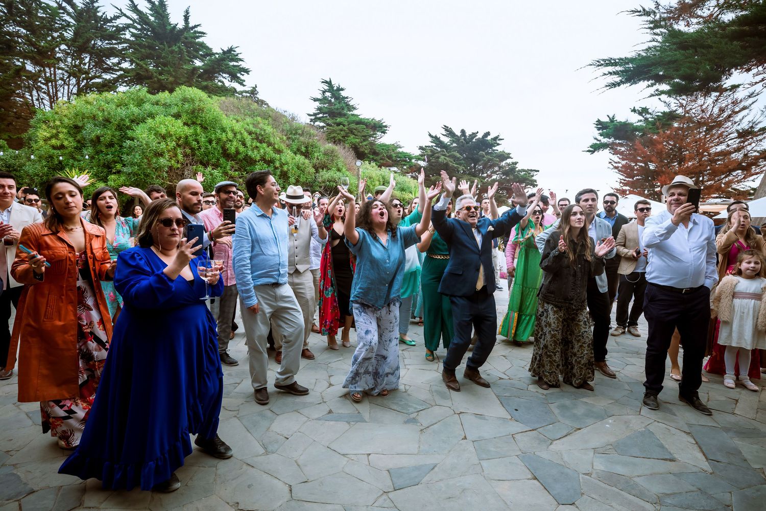Wedding guests dancing and celebrating outdoors at a coastal garden reception with lush greenery.