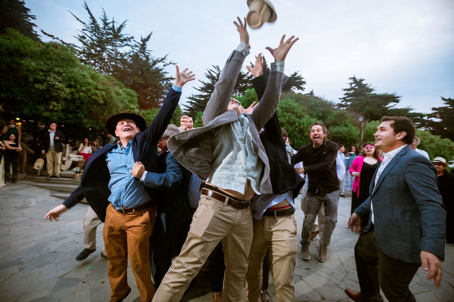 Men enthusiastically tossing hats in the air at an outdoor wedding celebration on a stone terrace.