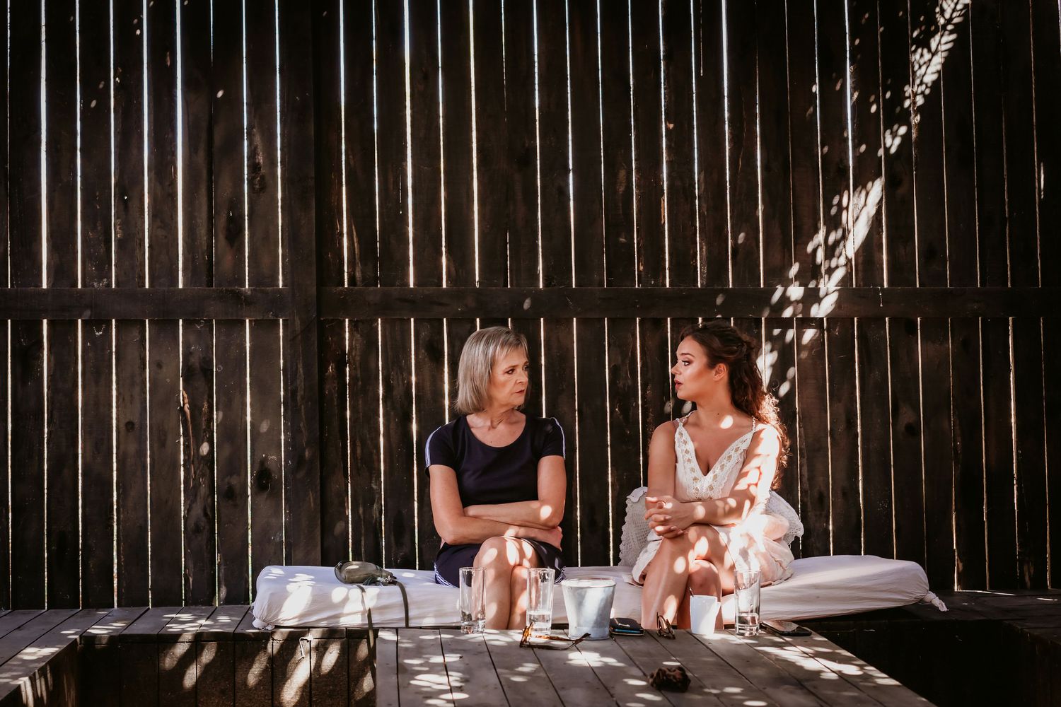 Two women sit together on a wooden deck in dappled sunlight, engaged in conversation near a rustic wooden wall.