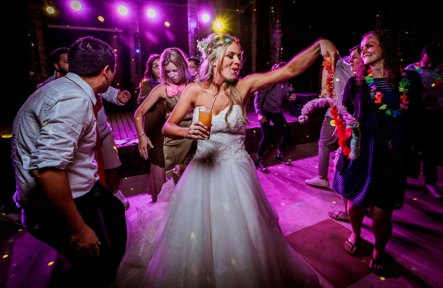 Bride in white ball gown dancing with guests under purple stage lights at reception.