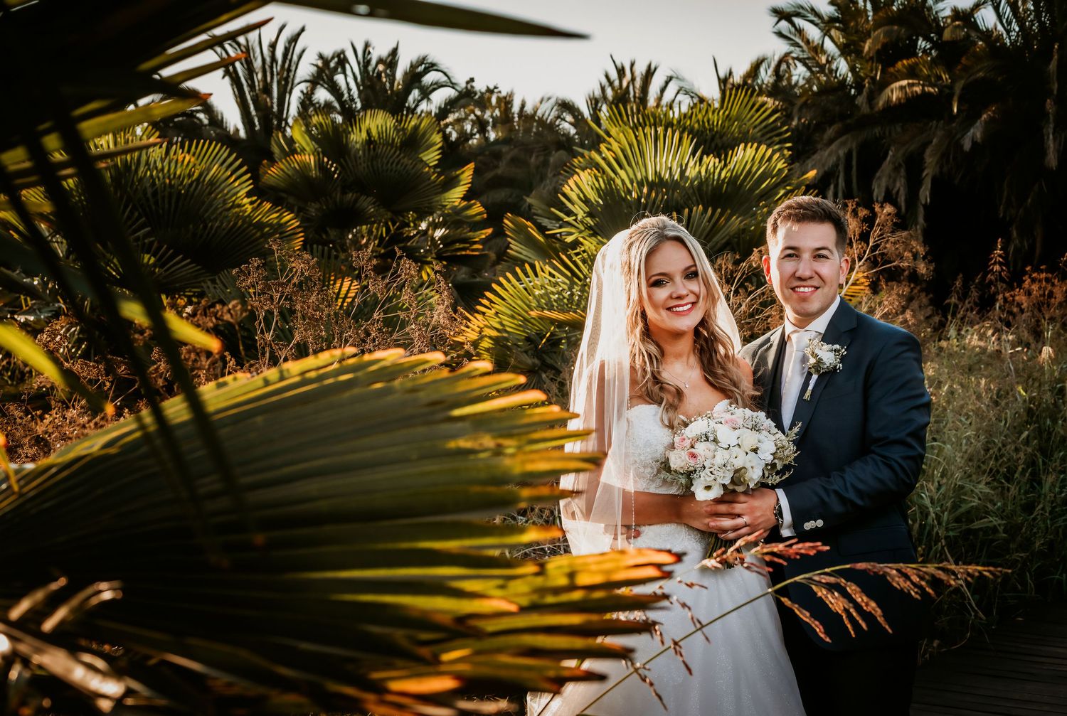Happy newlyweds smile together framed by lush tropical palm foliage in warm golden light.