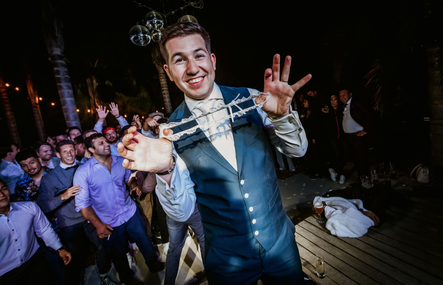 Smiling groom posing with hands raised surrounded by cheering wedding guests at outdoor reception.