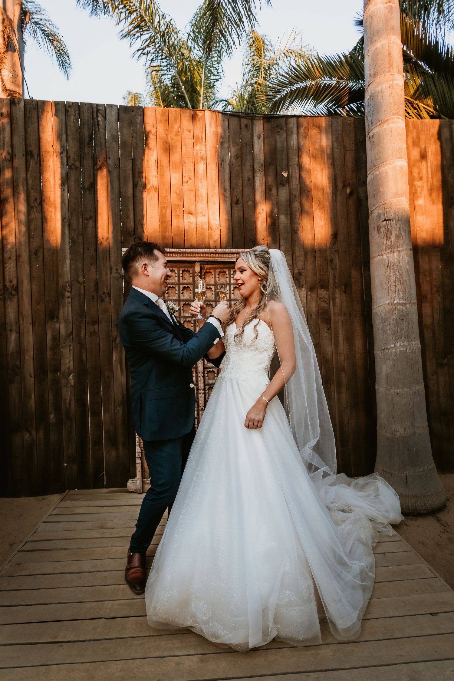 Bride and groom toast champagne glasses beside wooden fence and palm trees at golden hour.